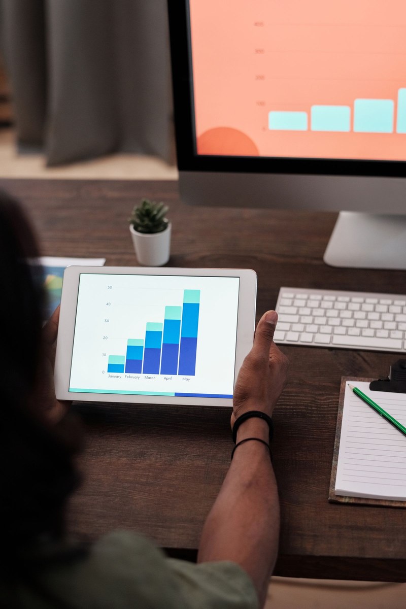 Hands holding a tablet showing a five-month stacked bar chart beside a keyboard and monitor